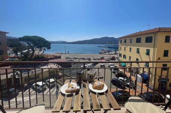 A balcony with wooden tables, two plates of food, and a view of a marina with boats and a clear blue sky.