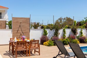 A poolside area with a wooden table and chairs, a pool, and a variety of plants and flowers. - Napa Bay View Villa 3 (Ayia Napa)