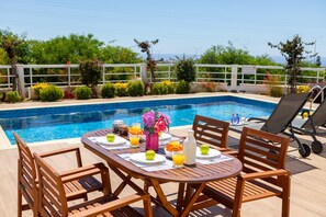 A poolside dining area with a table set for two, surrounded by wooden chairs, a pool, and a view of the sea. - Napa Bay View Villa 3 (Ayia Napa)