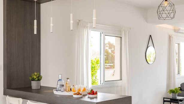 Una cocina moderna con una isla central, taburetes blancos de bar, iluminación colgante y vistas al exterior.
