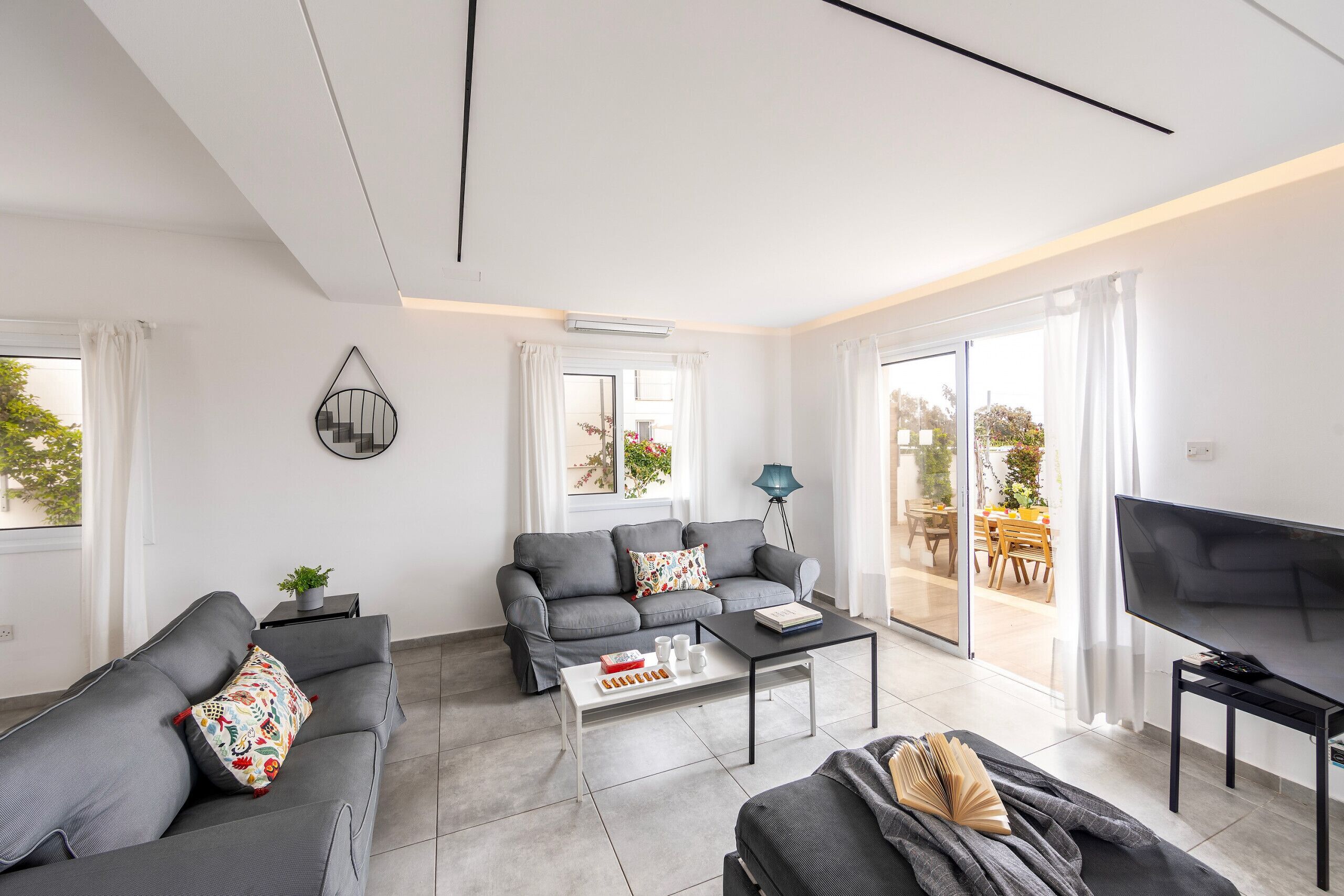 Una sala de estar moderna con sofás grises, una mesa de centro, un televisor y un ventanal corredero a un área exterior.