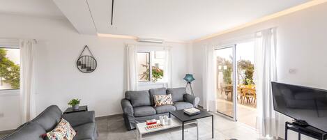 Una sala de estar moderna con sofás grises, una mesa de centro, un televisor y un ventanal corredero a un área exterior.