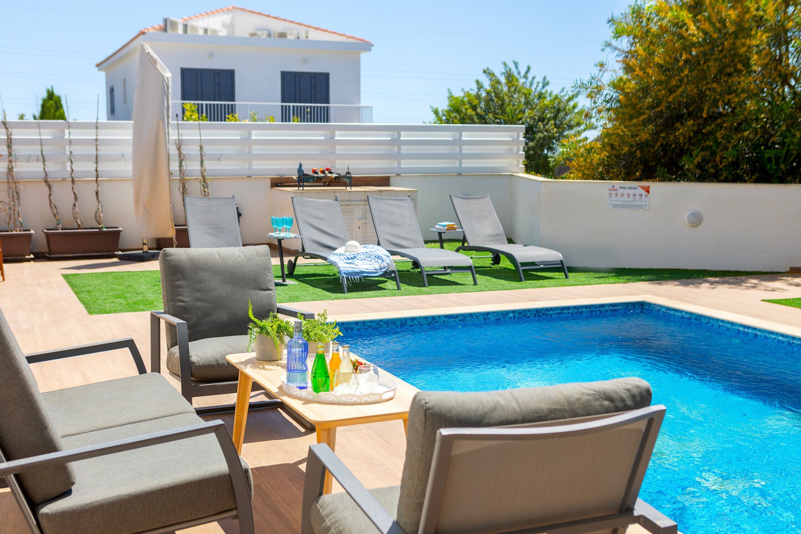 Un espace aménagé au bord de la piscine, avec des chaises longues, une table où sont servis des boissons, et un bâtiment blanc en arrière-plan.