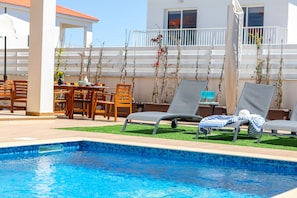 A swimming pool with lounge chairs, a table with chairs, and a white building in the background. - Napa Bay View Villa 7 (Ayia Napa)