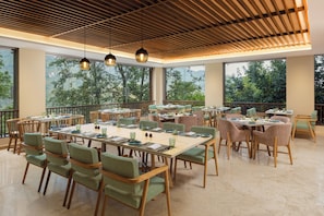 A spacious dining area with multiple tables and chairs, large windows offering views of greenery, and a wooden ceiling with hanging lights. - Fortune Resort & Wellness Spa Bhaktapur, Nepal - Member ITC Hotels' Group (Suryabinayak)