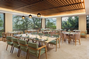 Un comedor espacioso con varias mesas y sillas, ventanas amplias que ofrecen vistas a zonas verdes y un techo de madera con luces colgantes.