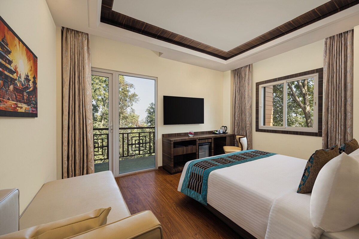 A hotel room with a bed, a desk, a television, and a balcony with a view of trees.