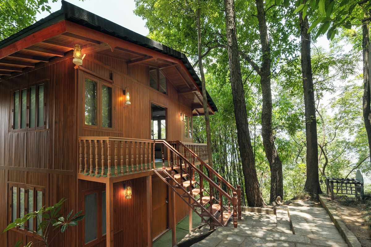A wooden cabin with a wooden staircase leading to a balcony, surrounded by trees.