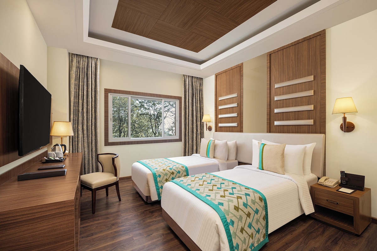 A hotel room with two beds, a desk with a flat-screen TV, a chair, a nightstand with a phone, and a window with curtains.