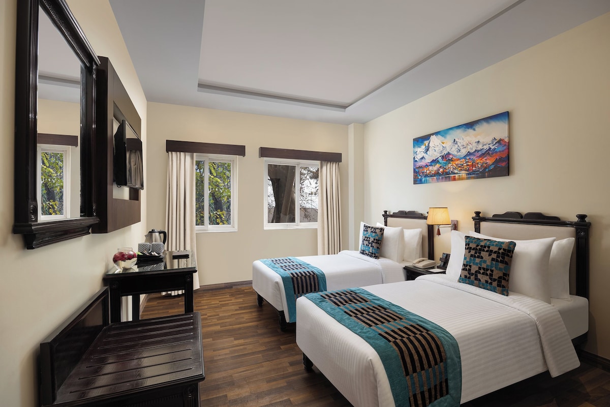 A hotel room with two beds, a desk, a mirror, a painting, and a window with curtains.