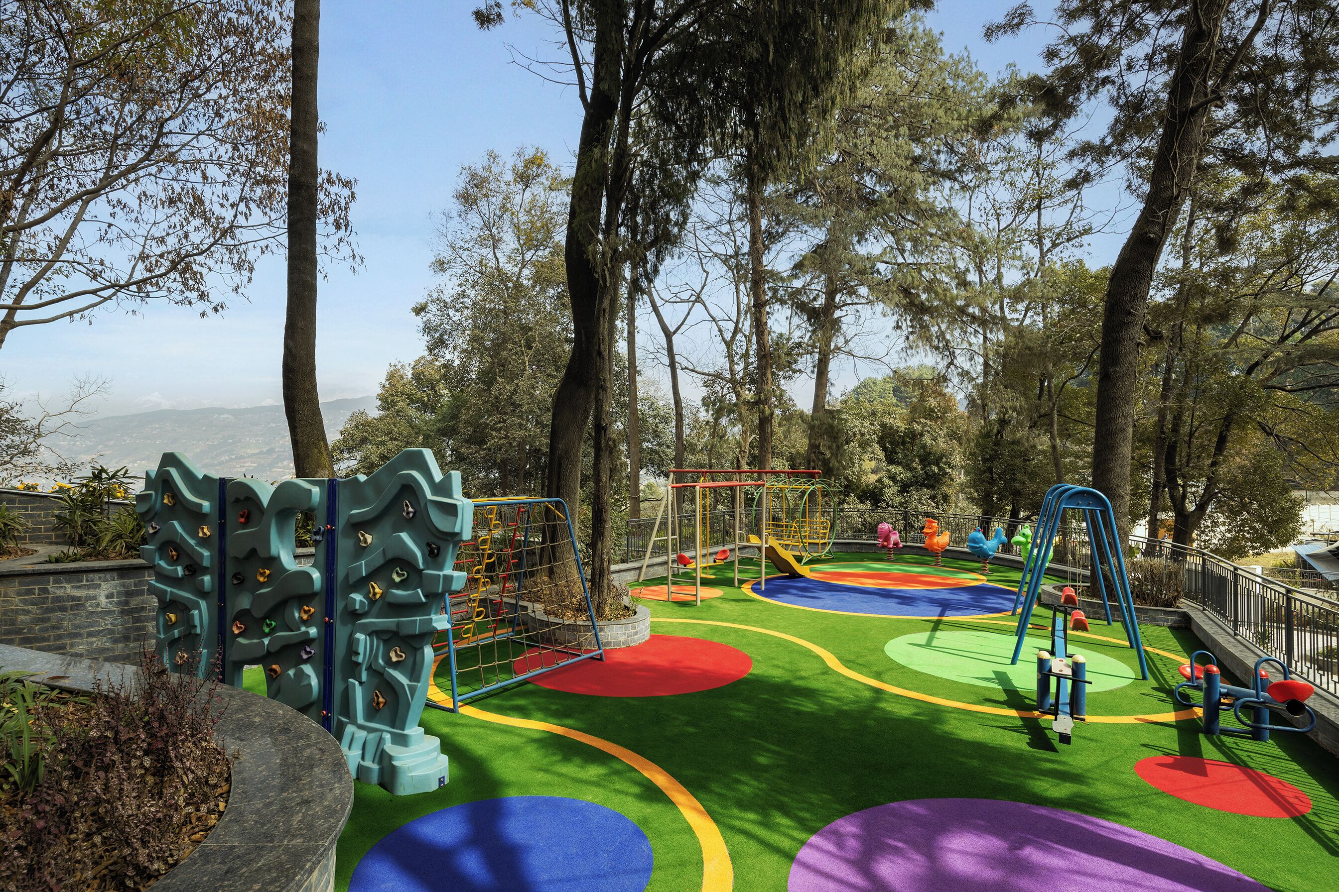A playground with climbing structures and colorful play areas, surrounded by trees and a view of the city.