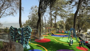 A playground with climbing structures and colorful play areas, surrounded by trees and a view of the city.