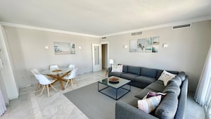 Una sala de estar moderna con un sofá seccional gris, una mesa de centro de vidrio y un comedor con sillas blancas.