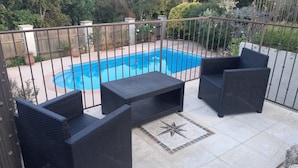 Eine Terrasse mit Schwimmbecken, umgeben von einem Metallzaun, mit Korbstühlen und einem kleinen Tisch.