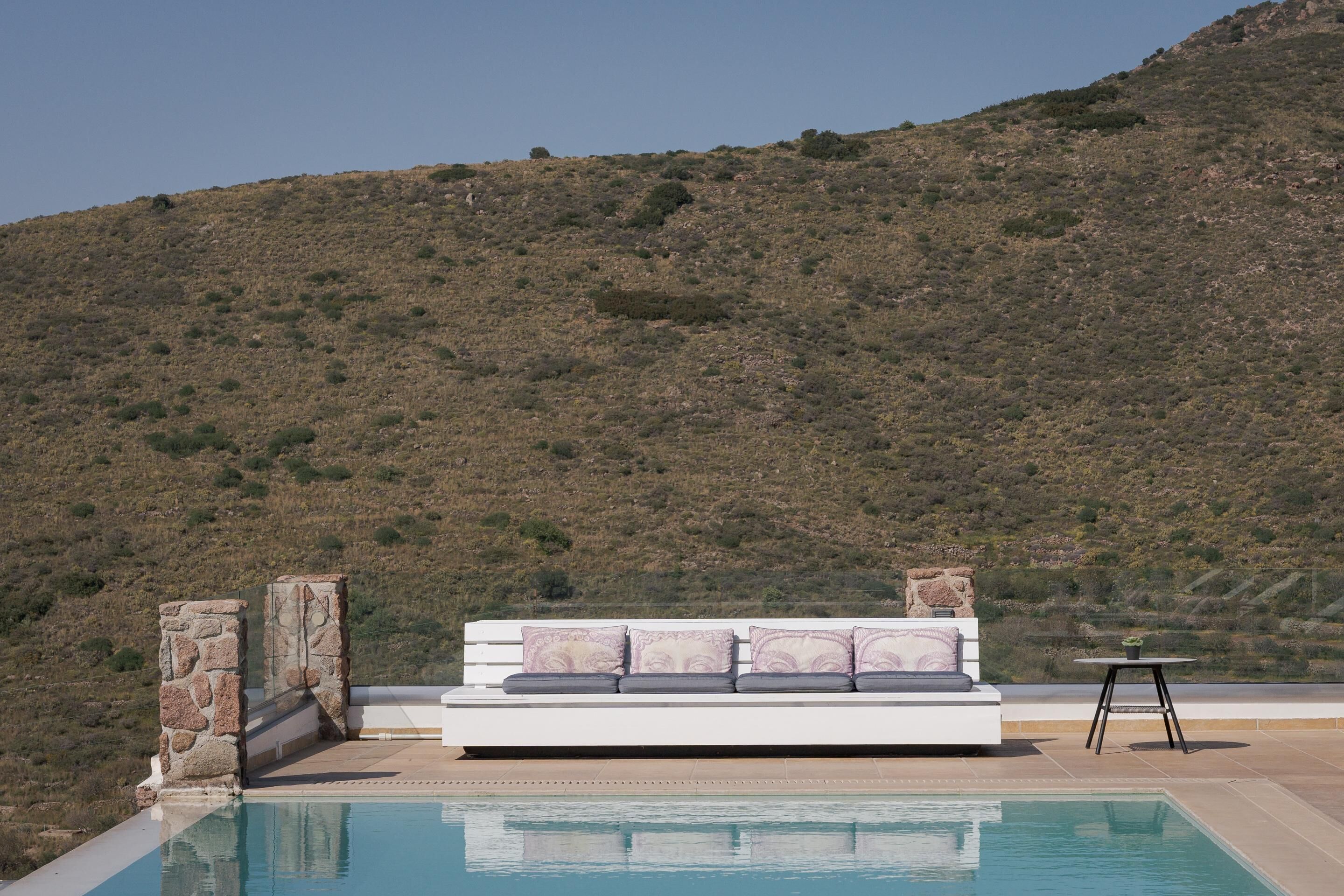 A poolside area with a white sofa, stone pillars, and a small table with a plant.