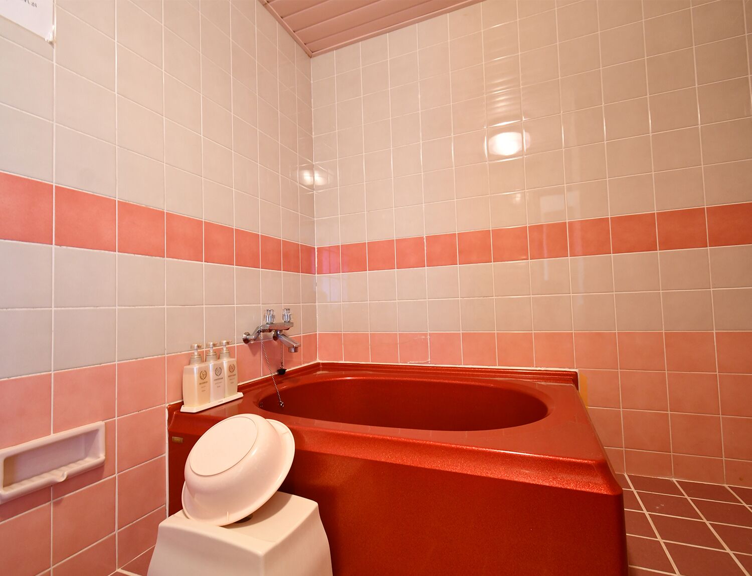 Un baño con una bañera roja, baldosas blancas y rosas, un dispensador de toallas blanco y un dispensador de jabón blanco.