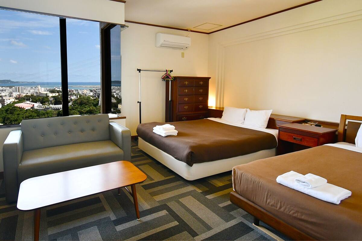 Quarto de hotel com duas camas, um sofá, uma escrivaninha e vista para a cidade.