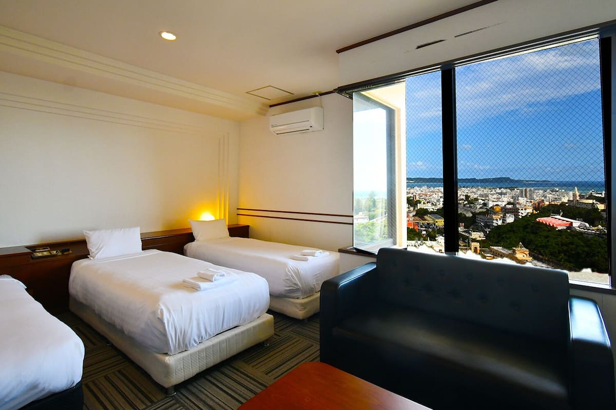 Quarto de hotel com duas camas, uma escrivaninha, uma cadeira e uma grande janela com vista para a cidade.