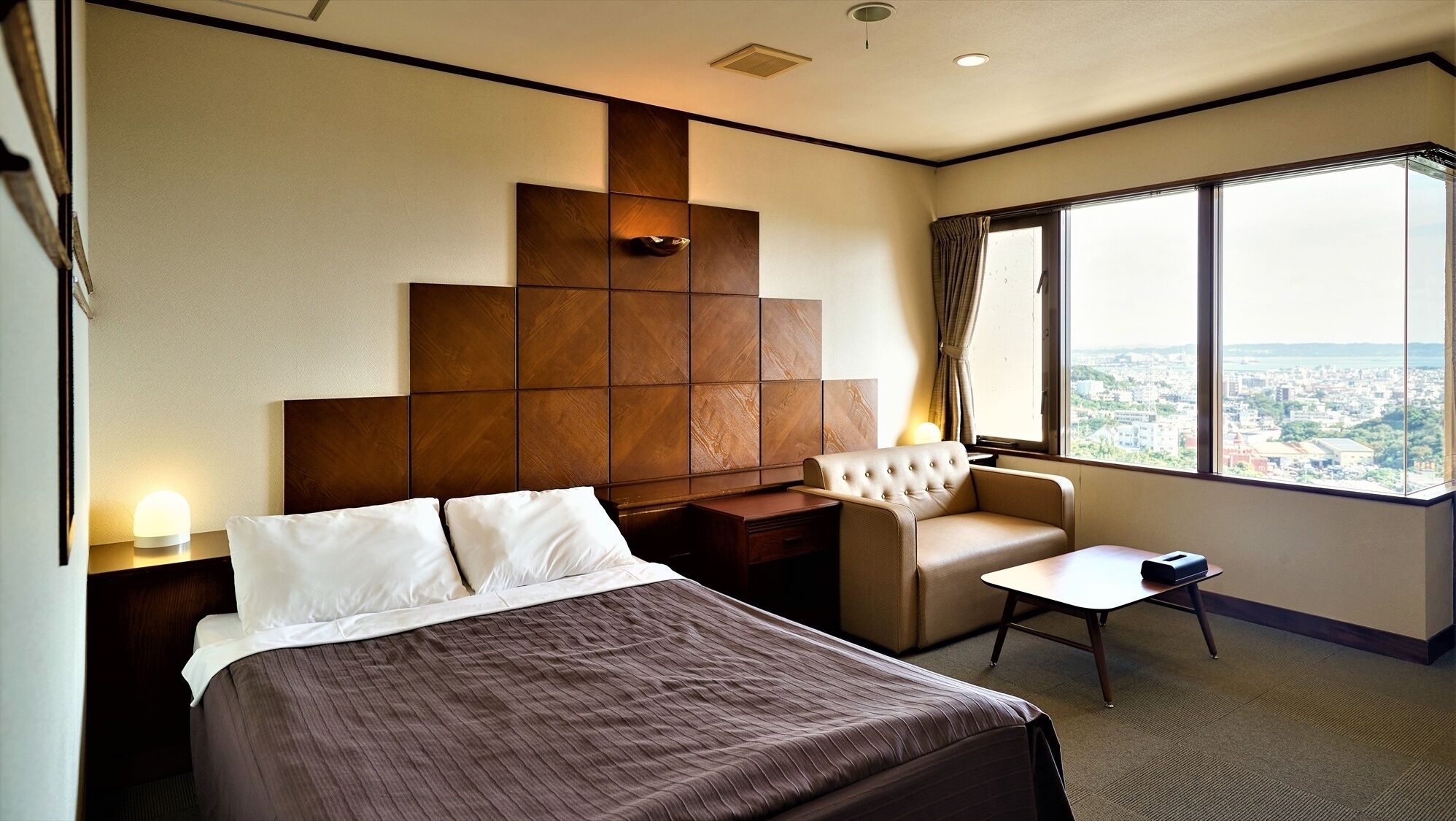 A hotel room with a bed, a small desk, a chair, and a view of the city through large windows.