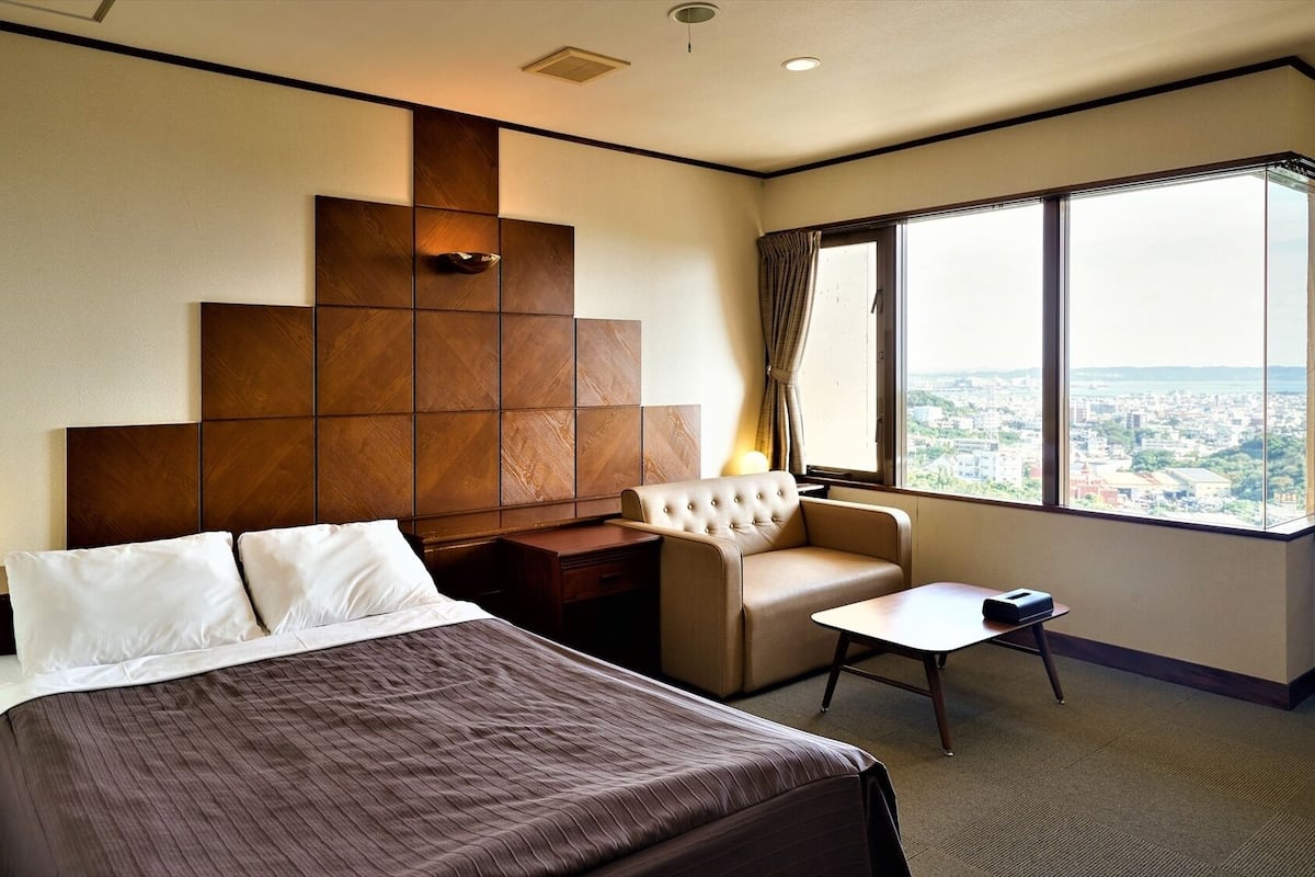 Quarto de hotel com uma cama, uma pequena escrivaninha, uma cadeira e vista da cidade através de grandes janelas.