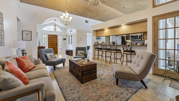 A spacious living room with a large sofa, a coffee table, and a chandelier. There is a kitchen area with bar stools and a refrigerator. - Beautiful River Front Home - Private Pool - Walk to Beach-Kayaks (Cocoa Beach)