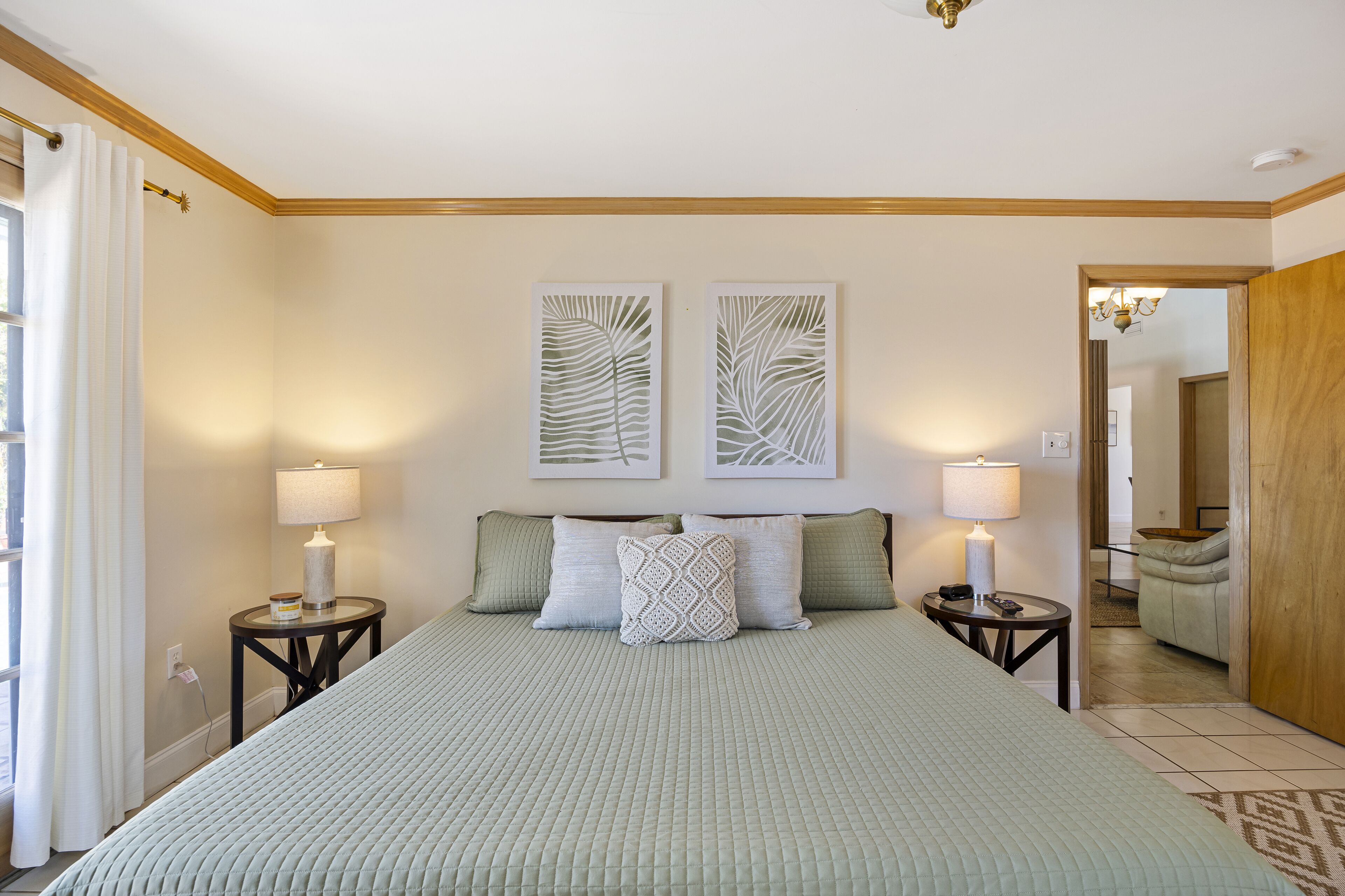 A bedroom with a large bed, two bedside tables with lamps, a window with curtains, and a door leading to another room.