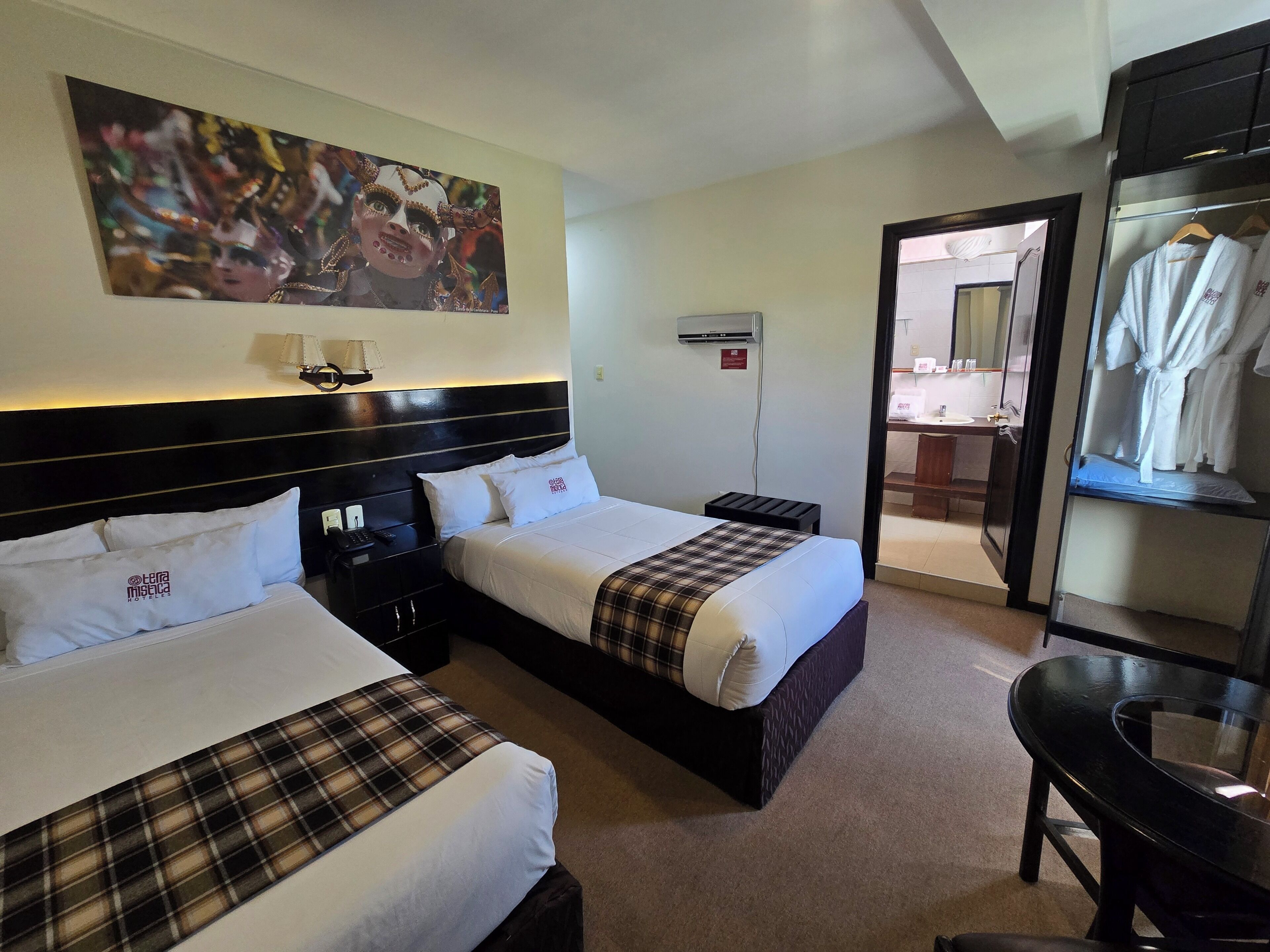 A hotel room with two beds, a desk, a chair, a painting, and a bathroom visible through an open door.