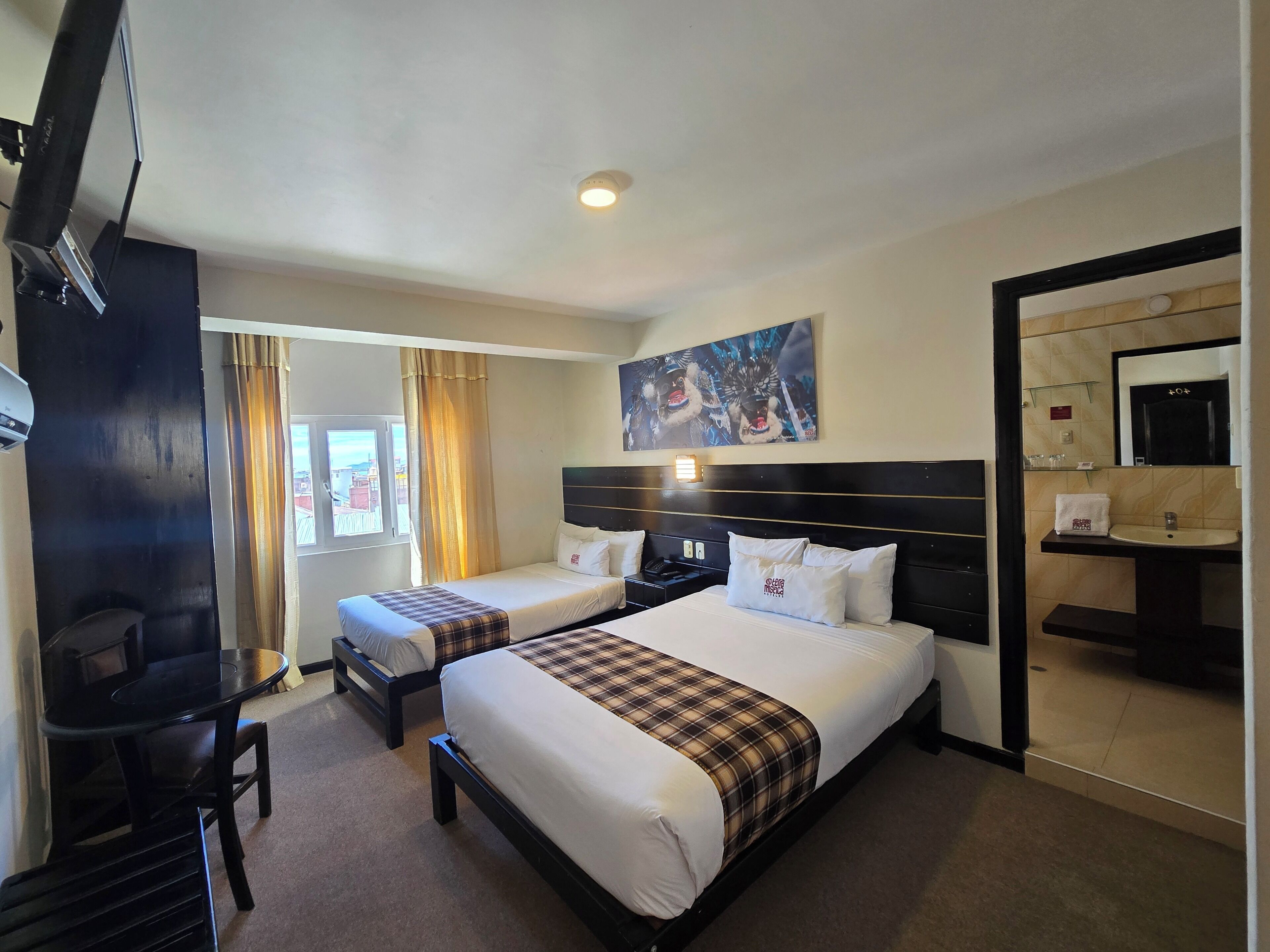 A hotel room with two beds, a desk, a chair, a painting on the wall, and a bathroom with a sink and mirror.