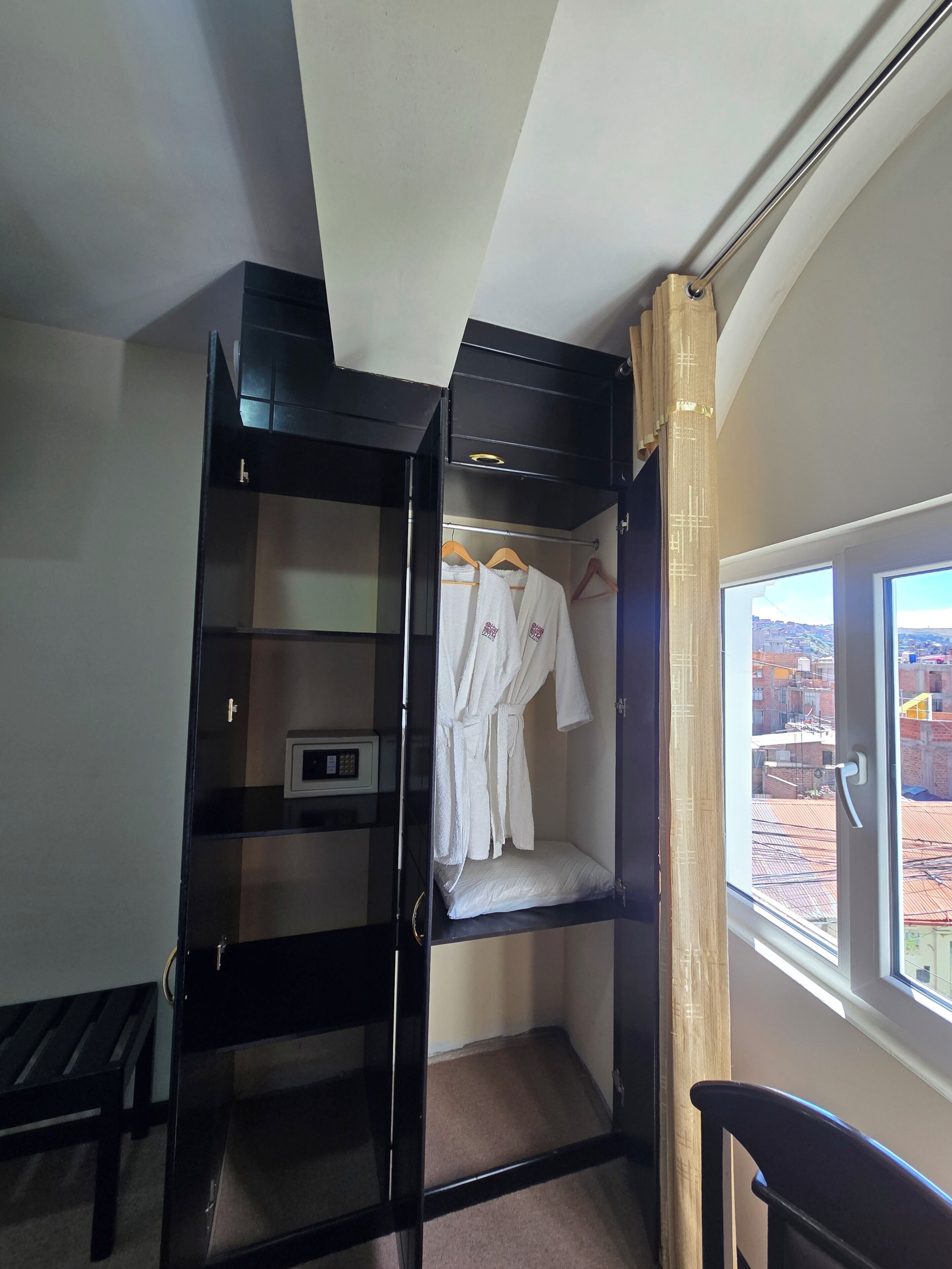 Habitación con armario negro, bata blanca colgada, una pequeña estantería con un dispositivo y ventana con vista a la ciudad.