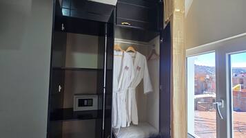 Habitación con armario negro, bata blanca colgada, una pequeña estantería con un dispositivo y ventana con vista a la ciudad.