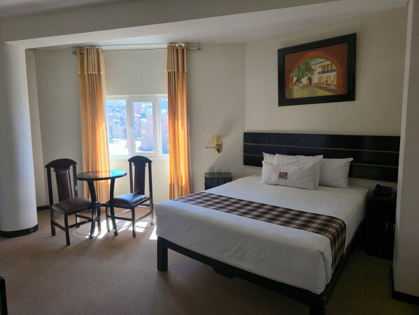 A hotel room with a bed, a table with two chairs, a window with curtains, a painting on the wall, and a nightstand. - TerraMistica - Puno Centro (Puno)