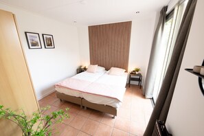 Quarto com cama grande, duas mesinhas de cabeceira, cabeceira de madeira e janela com cortinas.