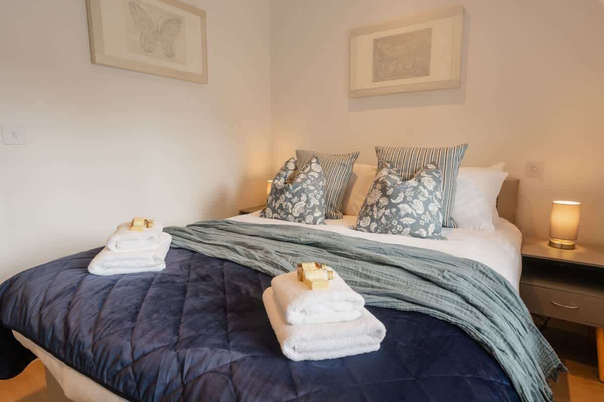 Una cama bien hecha con un colcha azul, dos almohadas con motivos florales y una mesita de noche con una lámpara.