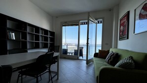 A modern living room with a dining area, a green sofa, a bookshelf, and a large sliding glass door leading to a balcony with a city view. - TORRE AZZURRA - SUNSET (Follonica)