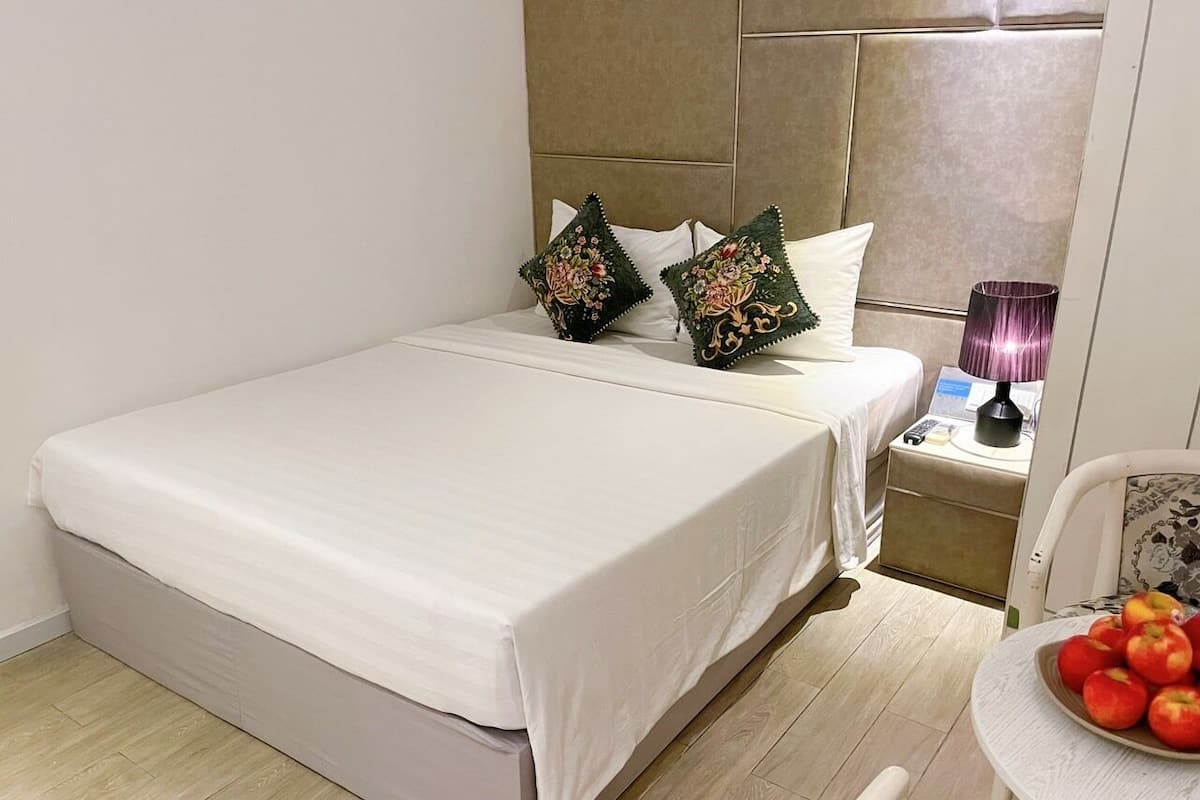 A neatly made bed with white linens, two decorative pillows, a bedside table with a lamp, and a small table with a bowl of fruit.