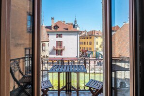 Ein Balkon mit Tisch und zwei Stühlen, der auf eine Straße mit Gebäuden und einem klaren Himmel blickt.