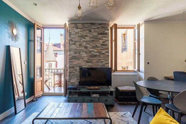 A living room with a stone wall, a TV on a wooden stand, a dining table with chairs, and a balcony visible through an open door.