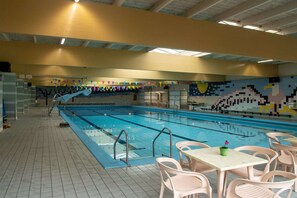 Una piscina interior con suelo de baldosas, sillas junto a la piscina y un mural colorido en la pared.
