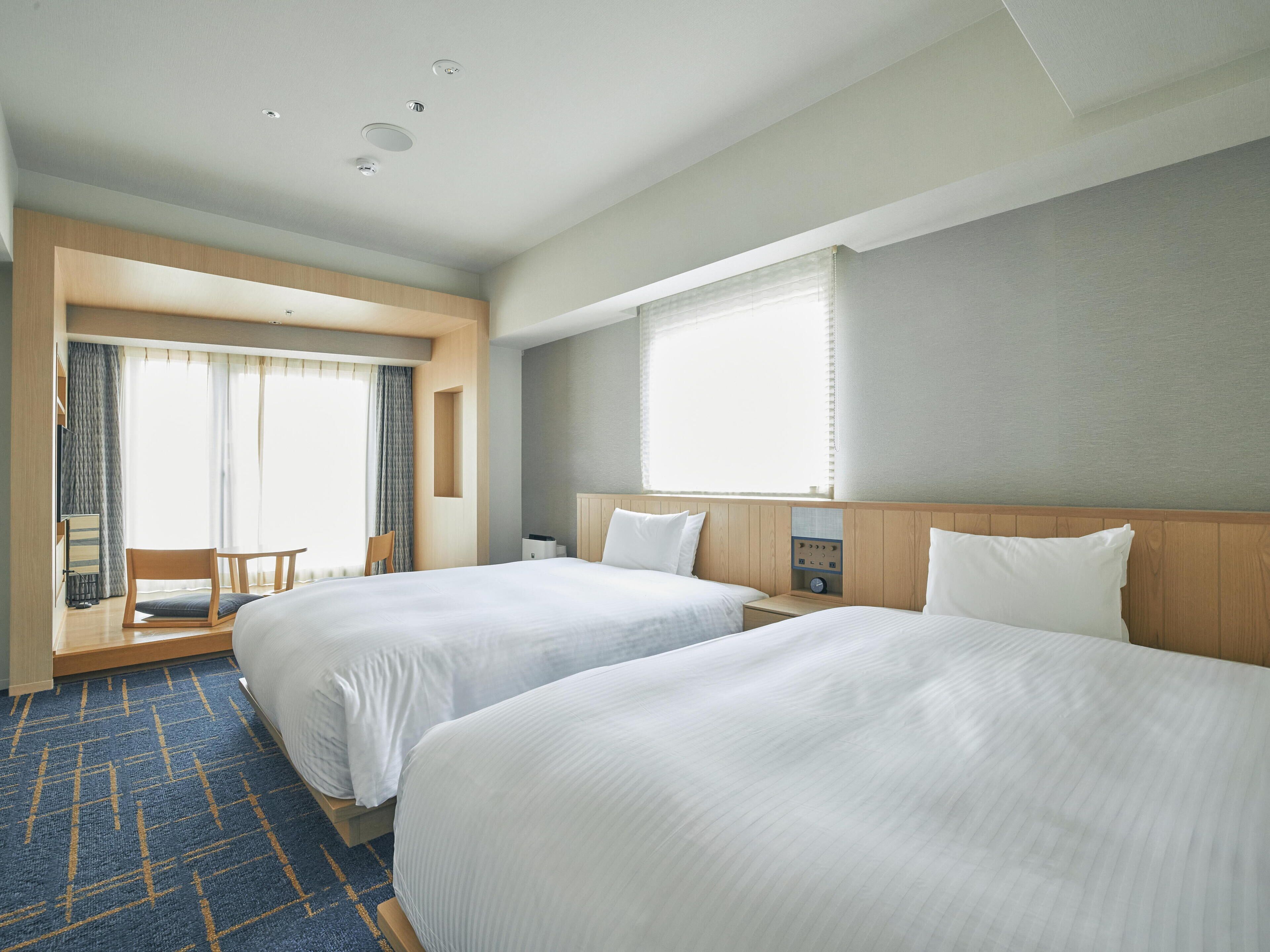 Habitación de hotel con dos camas, una zona de estar pequeña con una silla y una mesa, un ventanal grande con cortinas y un panel de control en la pared.