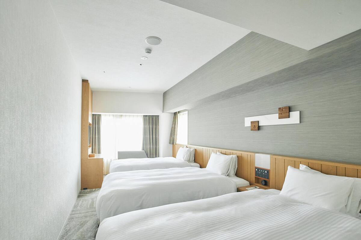 A hotel room with two beds, a wooden headboard, a bedside table with a clock, and a large window with curtains.