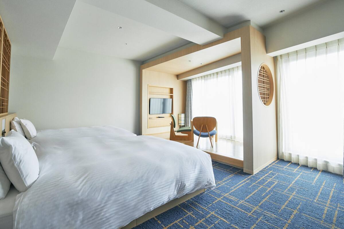 A modern hotel room with a large bed, a desk with a chair, a television, and a large window with curtains.