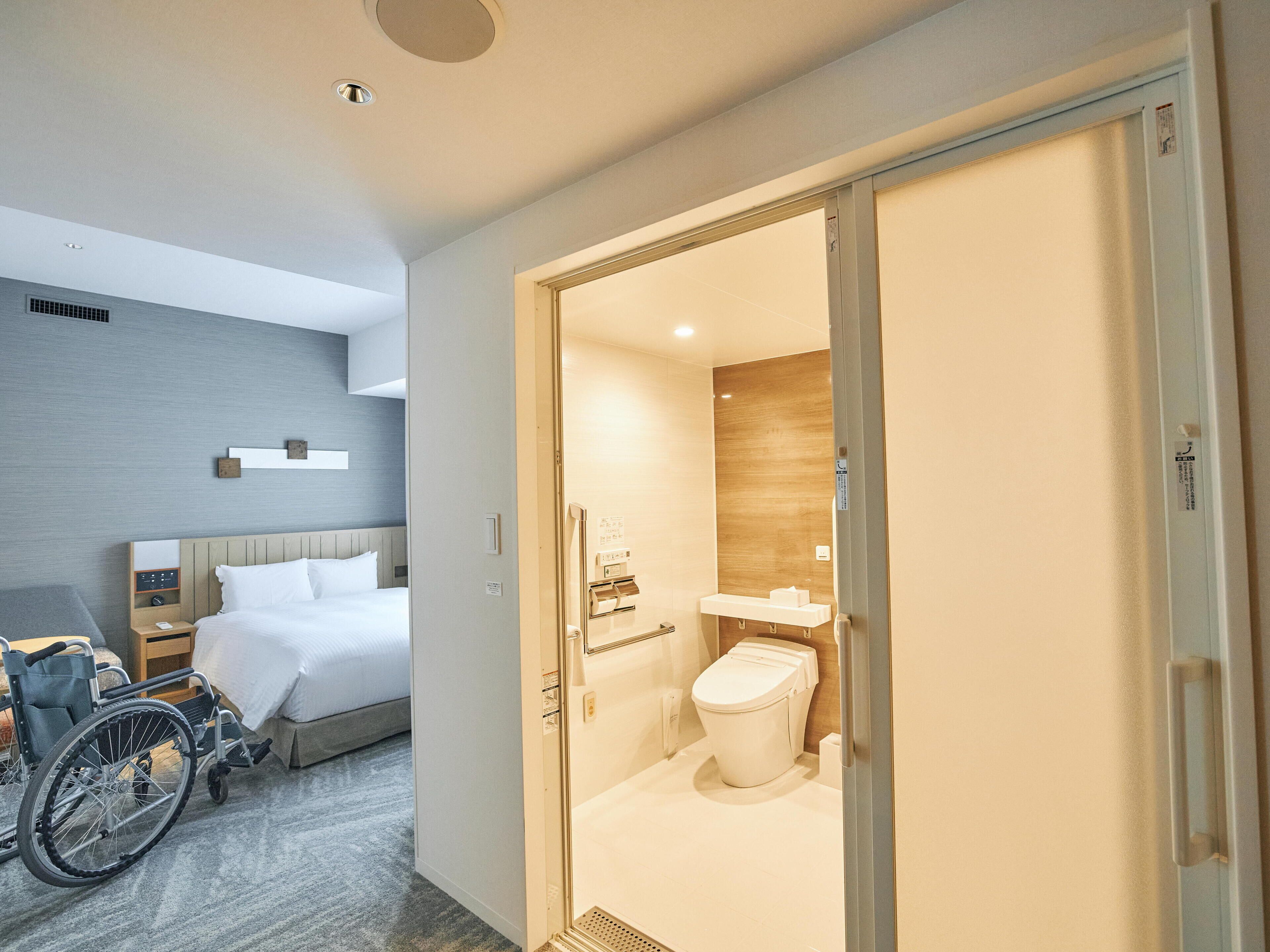 Ein Hotelzimmer mit Rollstuhl, einem Bett, einem Nachttisch und einem Badezimmer mit Toilette und Waschbecken.