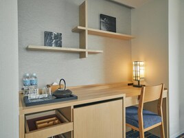 A hotel room with a wooden desk, a chair, a lamp, and two shelves with artwork.