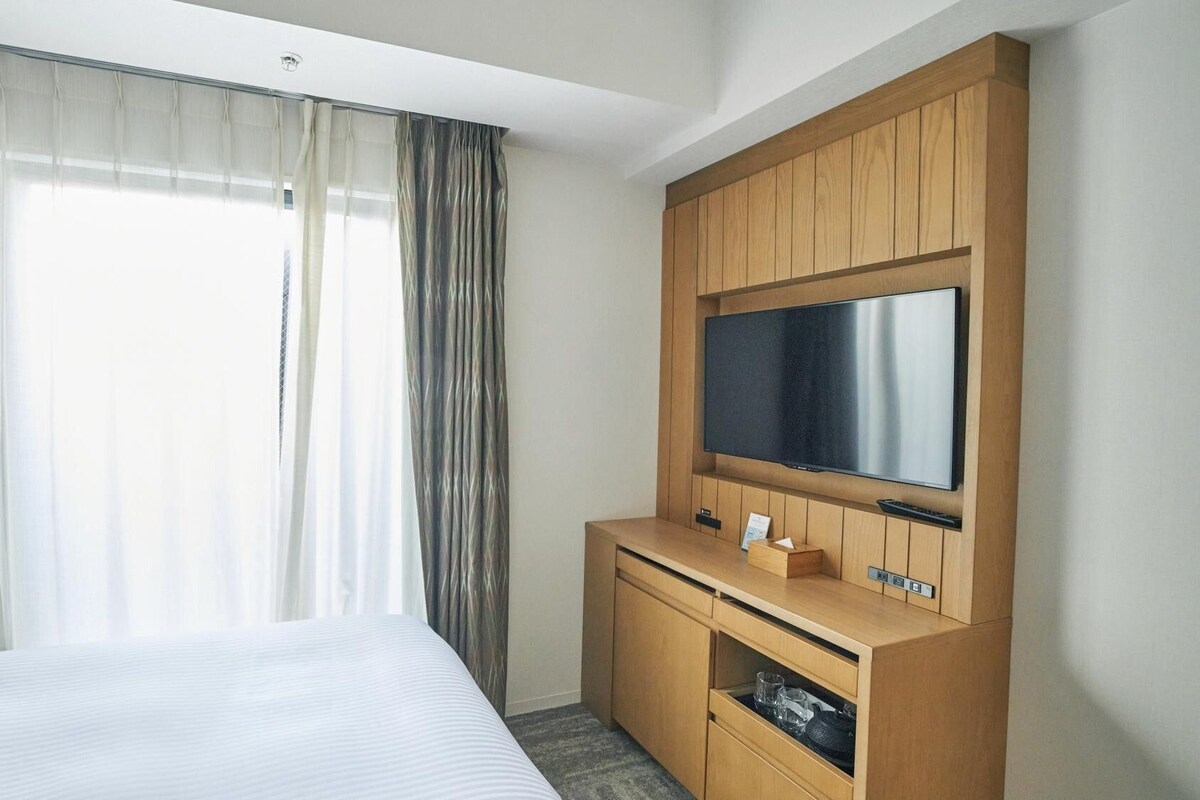 Une chambre d’hôtel avec un lit, un meuble en bois pour la télévision et une grande fenêtre avec des rideaux.