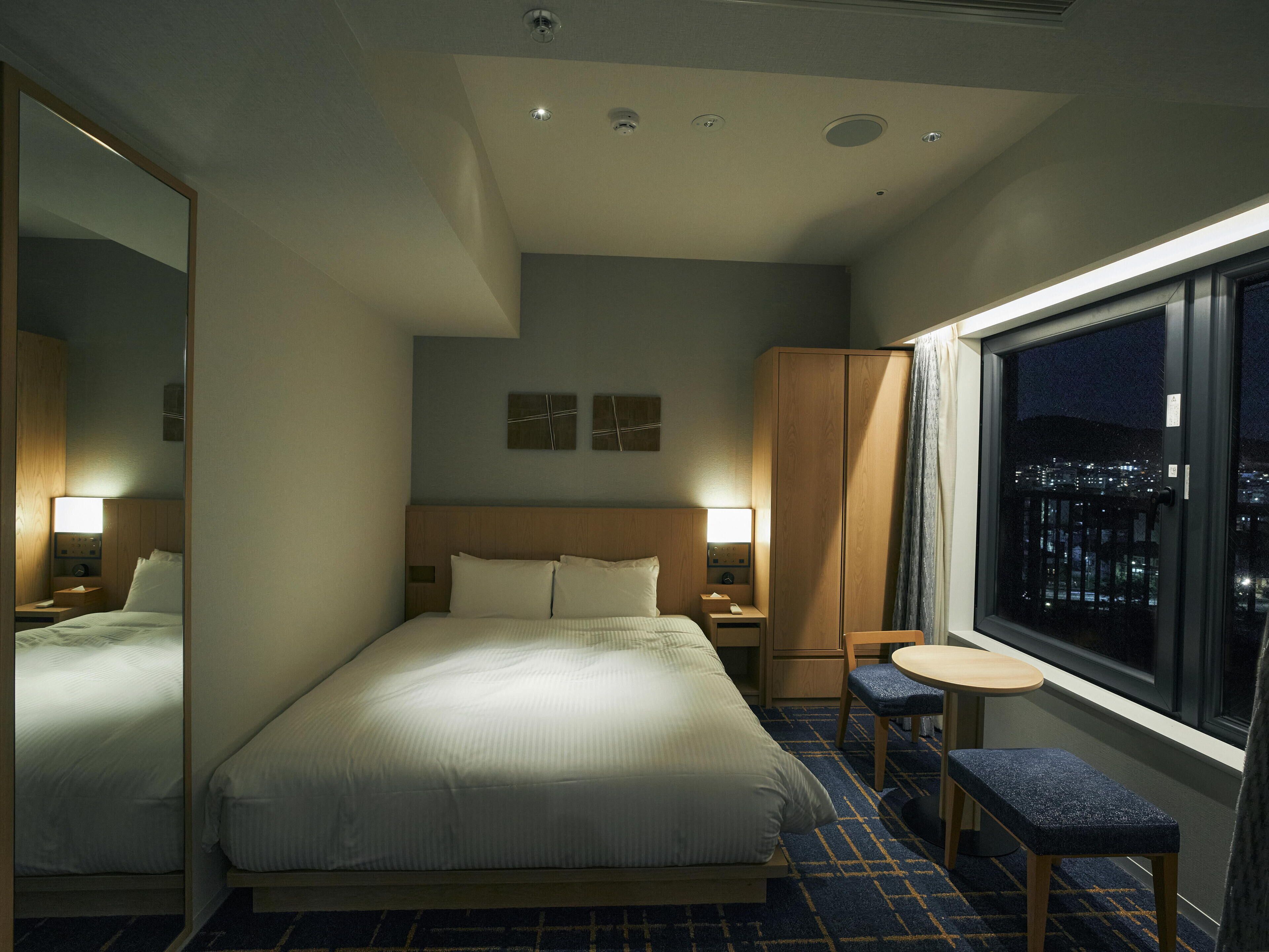 Une chambre d’hôtel avec un grand lit, une petite table, une chaise, une armoire et une fenêtre donnant sur la ville.