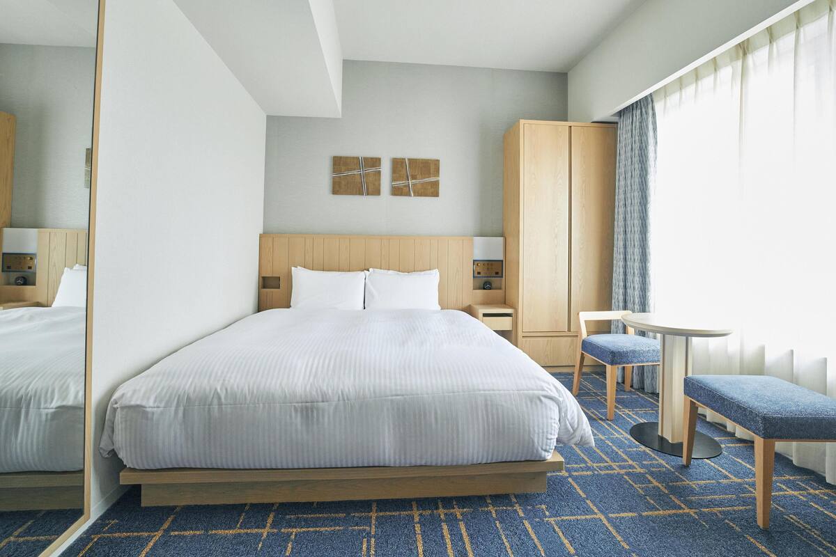 A hotel room with a large bed, a wooden wardrobe, a small table, and a bench.