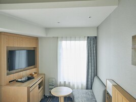 A compact hotel room with a flat-screen TV mounted on a wooden cabinet, a small round table, a sofa, and a window with sheer curtains.