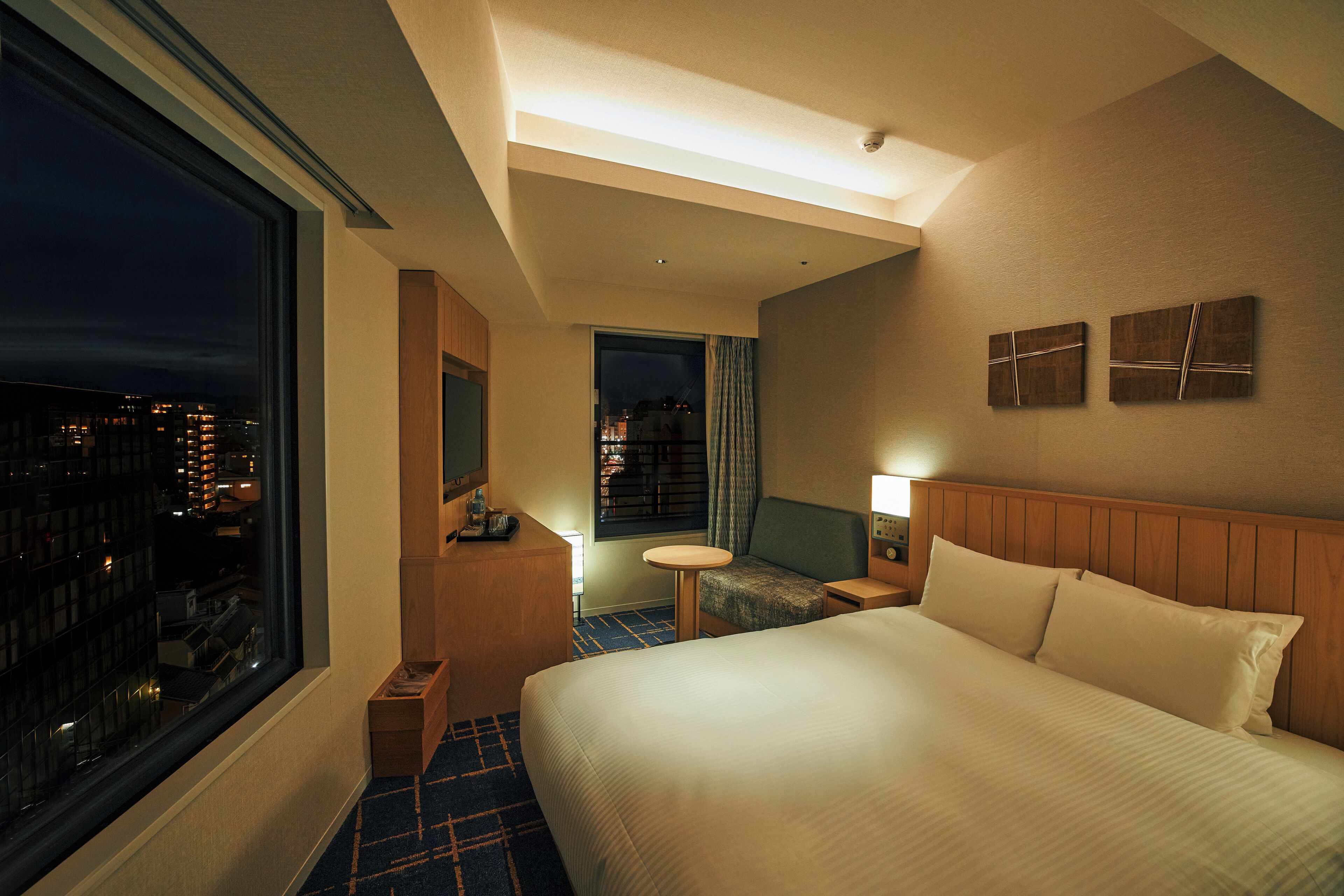 A hotel room with a large bed, a small table, a chair, a nightstand, a window with a city view, and a wall-mounted artwork.