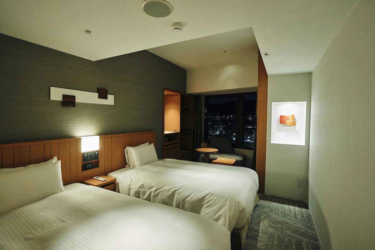 A hotel room with two beds, a nightstand, a small table, a chair, a window with a night view, and a wall-mounted artwork.