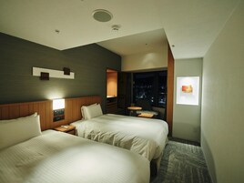 A hotel room with two beds, a nightstand, a small table, a chair, a window with a night view, and a wall-mounted artwork.